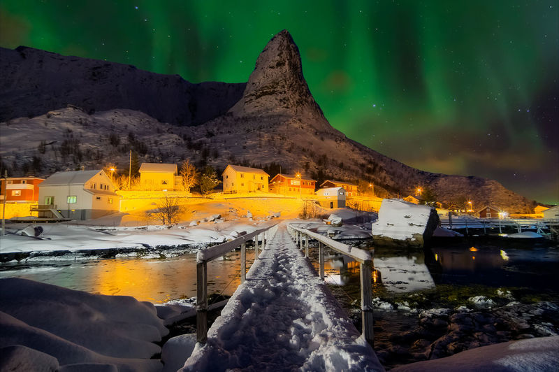 Wooden bridge over river leading towards illuminated houses and mountain at winter