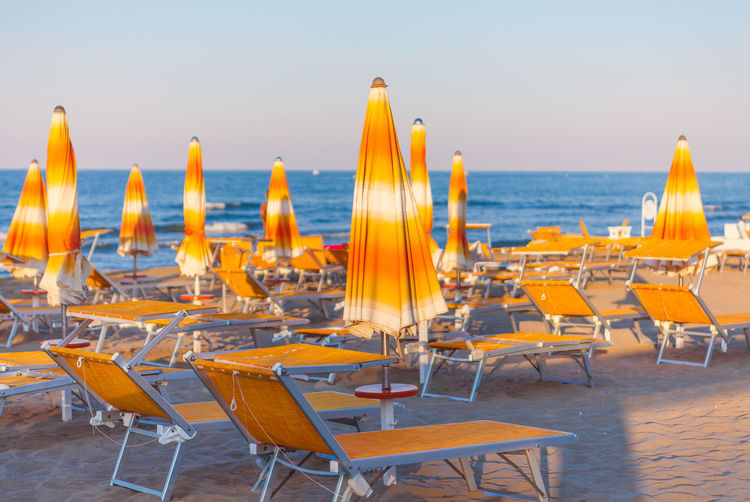 Chairs with tables and parasols arranged at beach against sky during sunset