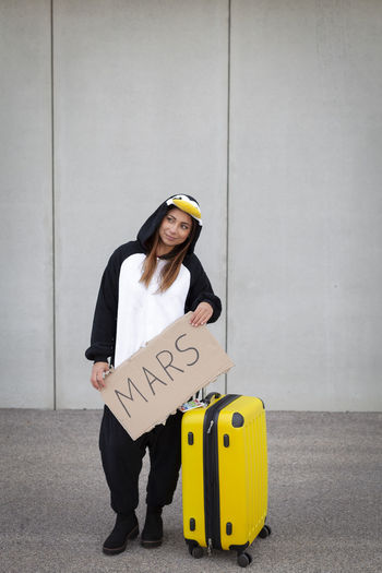 Woman holding text written on cardboard while standing by luggage against wall