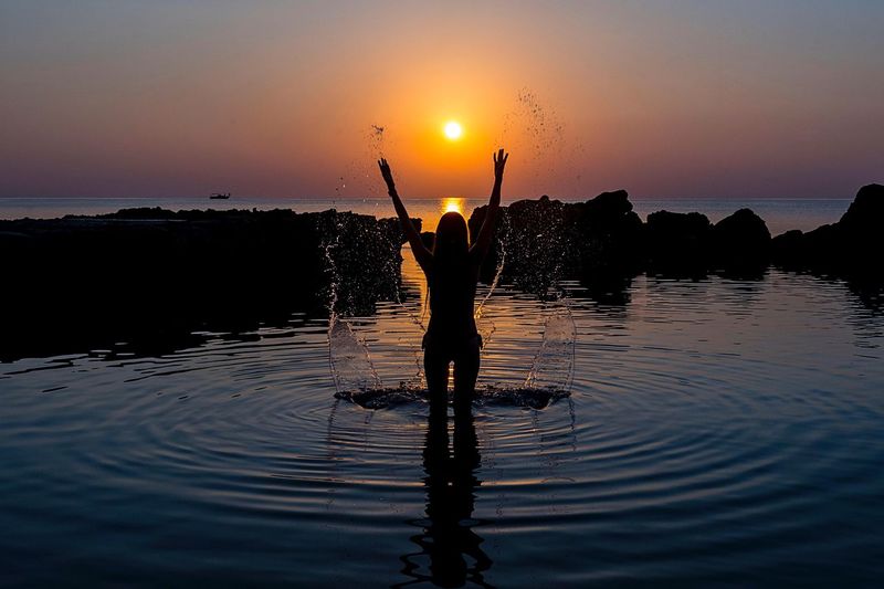 Rear view of silhouette woman splashing water in sea against sky during sunset