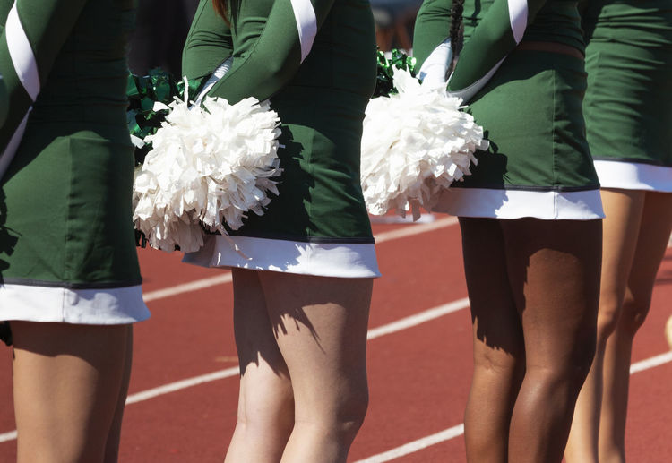 Close up of cheerleaders in green uniforms holding white pom poms behind backs.