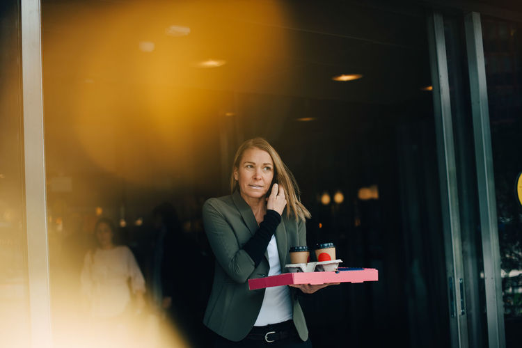 Mature businesswoman talking on smart phone while holding food and drink against cafe