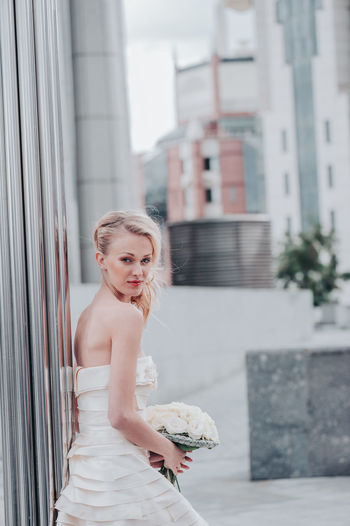 Beautiful bride with bouquet against wall in city