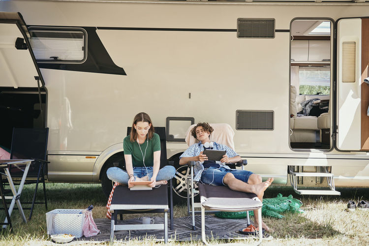 Teenage siblings watching movie on digital tablet while sitting on folding chairs against motor home at campsite