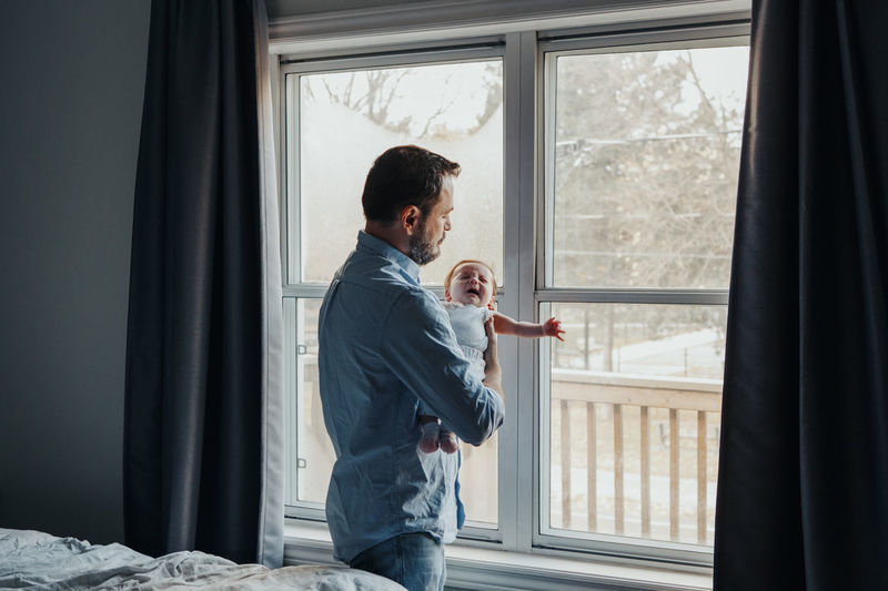 Father carrying baby daughter while standing by window at home