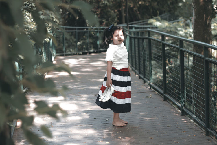 Full length portrait of girl holding shoe while standing on footbridge