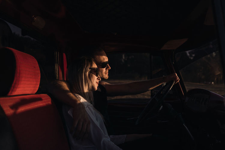 Side view of loving couple in sunglasses embracing in vintage car while riding in nature in evening