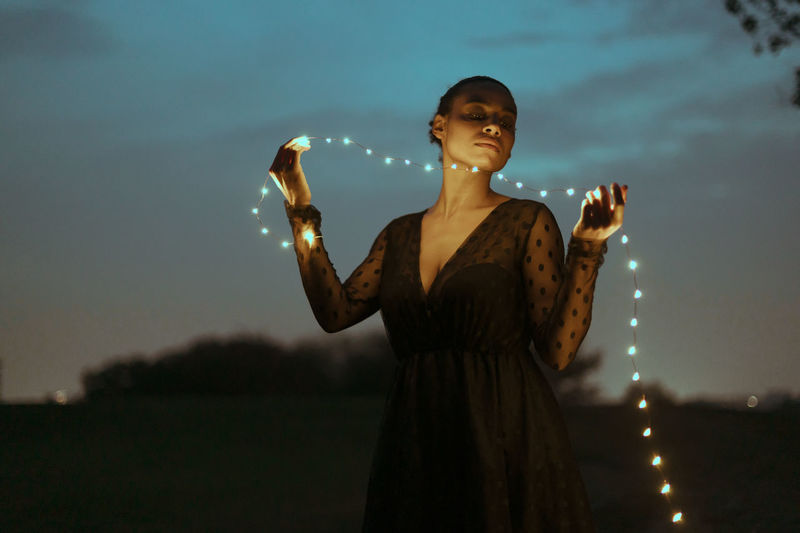 Beautiful woman with holding string lights outdoors at dusk