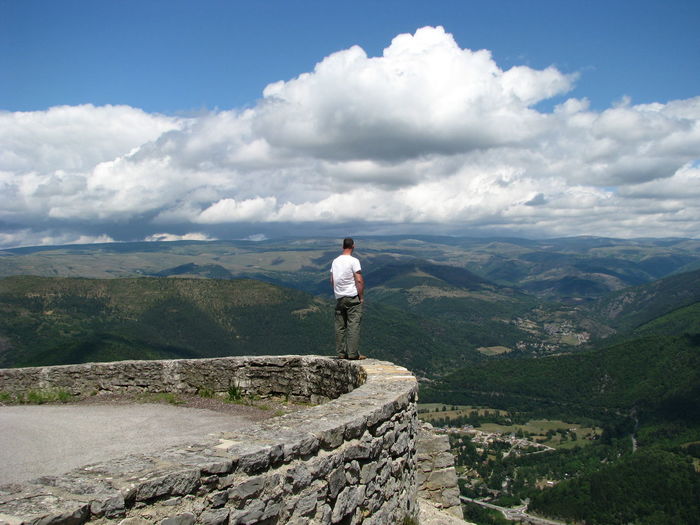 Rear view of man standing on surrounding wall by mountains against sky