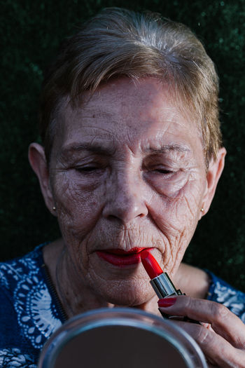Old woman painting her lips in red looking at herself in the mirror