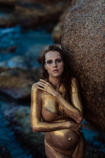 A pregnant girl in gold glitter stands against the backdrop of the ocean and stones