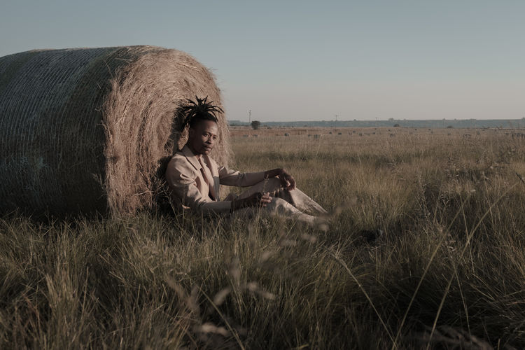 Young man sitting by hay bale at agricultural landscape