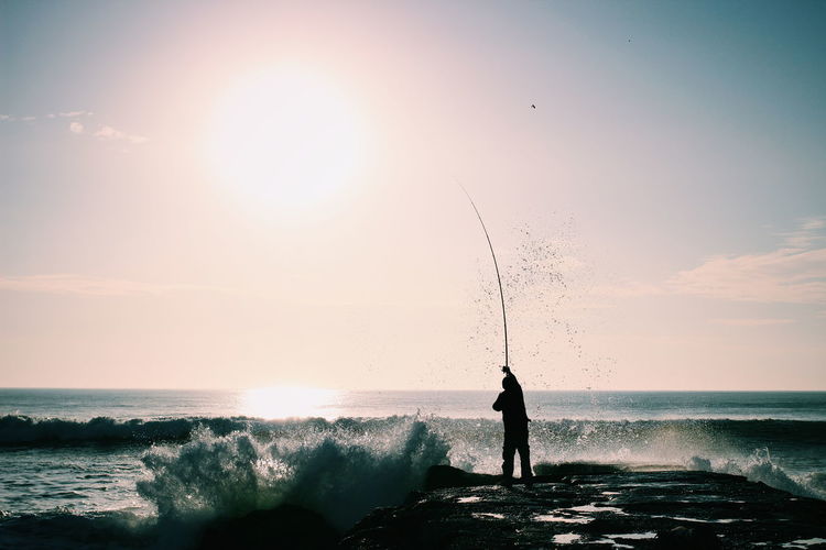 Rear view of man fishing in sea against sky on sunny day