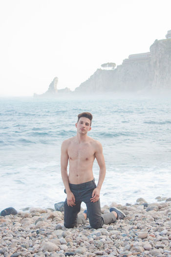 Shirtless young man kneeling on stones at beach