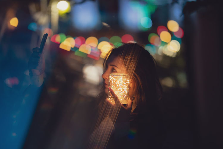 Young woman holding illuminated jar and mobile phone at night