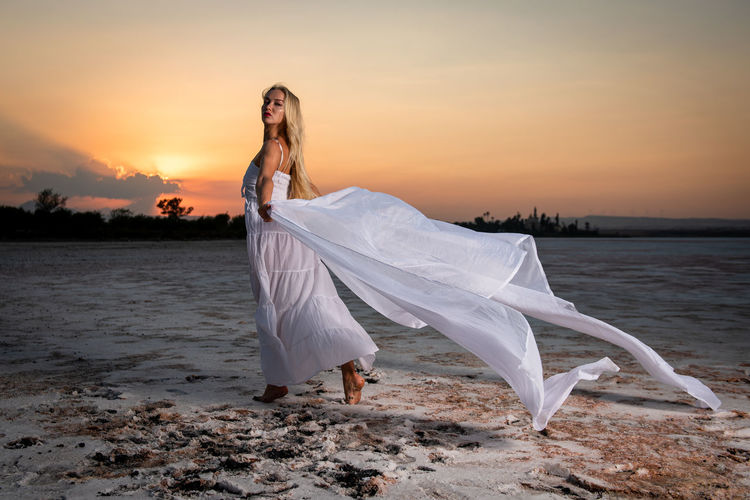 Portrait of beautiful woman standing at beach against sky during sunset