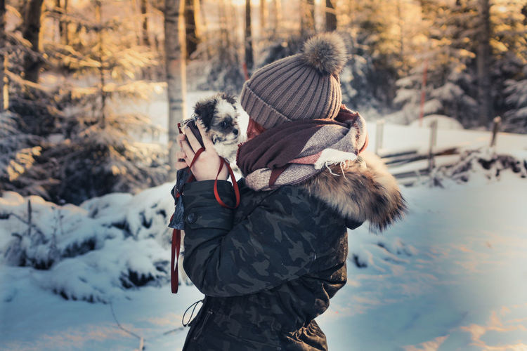 Side view of woman carrying puppy while standing on snow in forest during winter