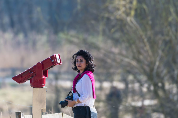 Portrait of woman holding camera while standing by hand-held telescope