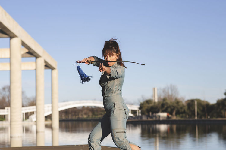 Confident woman athlete practicing martial arts with sword against lake at park