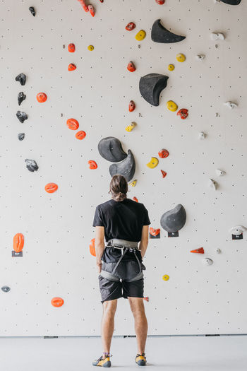 Full body back view of anonymous sportive male alpinist with safety harness in activewear standing near artificial climbing wall in gym