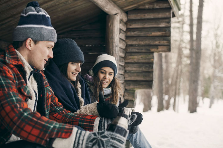 Man giving drink to female friends while sitting at log cabin during winter