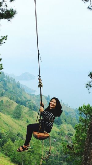 Portrait of cheerful woman on swing over mountain against sea