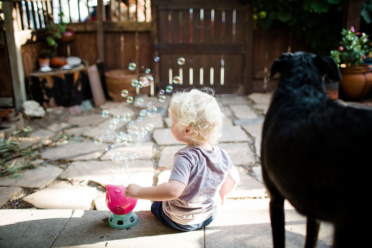 Blond boy playing with bubbles while black dog looks around yard