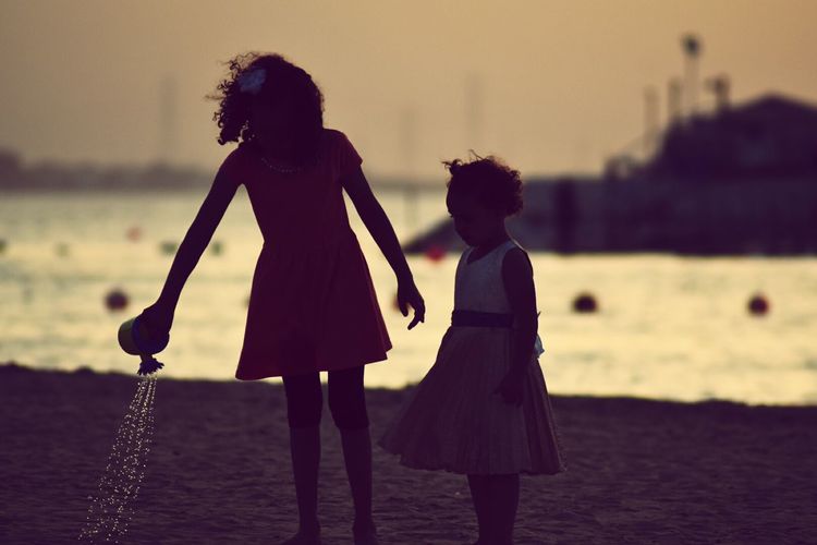 Silhouette children standing at beach against sky during sunset
