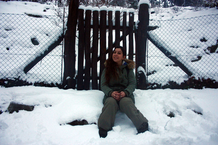 Girl in winter outfit sitting in front of a wooden fence among the white snow in daylight in tuscany