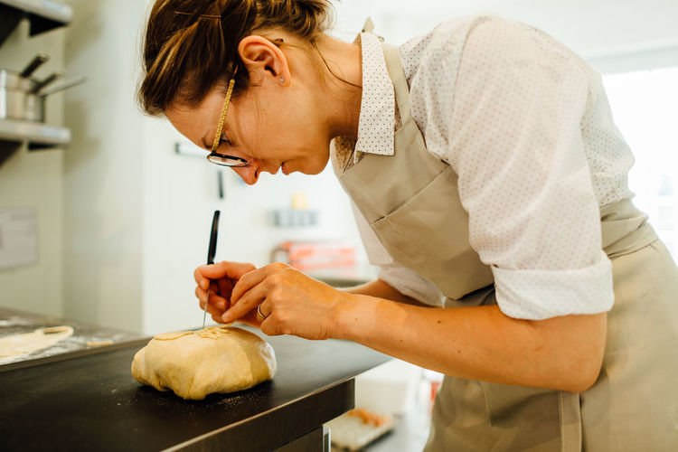 Female baker in uniform is decorating bread while working in bakery