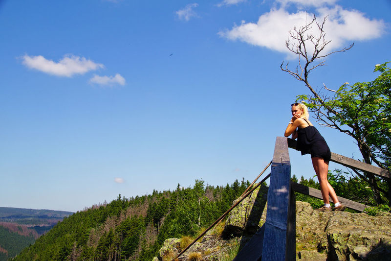 Low angle view of young woman standing on mountain against blue sky during sunny day