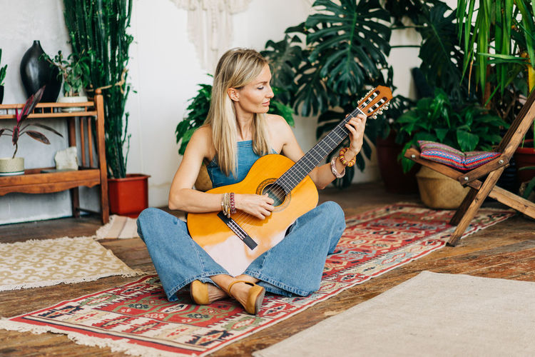 Young blond woman musician playing guitar sitting on floor in home greenhouse