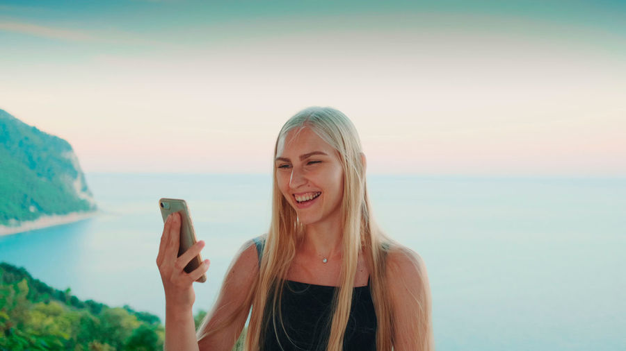 Smiling young woman using smart phone against sea