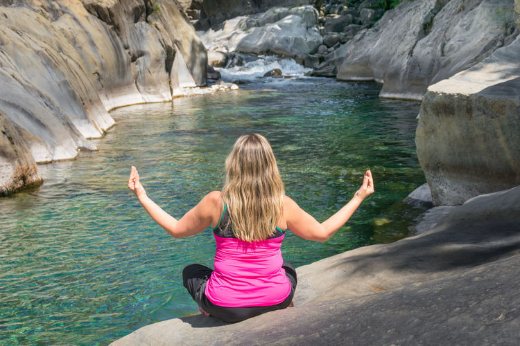 Woman backwards wearing sporty clothes practicing relaxing yoga exercise in an idyllic place next to a river and a waterfall between white rocks. the yogi is sitting on the floor with arms up