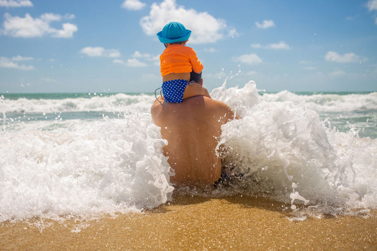 Father's day. dad protect. the baby sits on dad shoulders, dad sits on the beach and a wave comes.