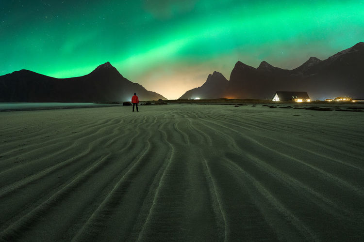 Back view of unrecognizable male tourist admiring snowy mounts under bright aurora borealis lights on lofoten island