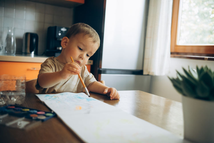 Child draws with a brush and paint in an album sitting at the table