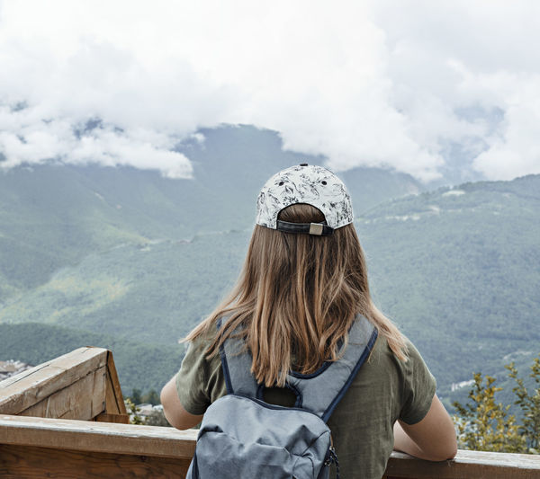 Rear view of young blonde woman in cap looking at view of caucasian mountains and cloudy sky in hike