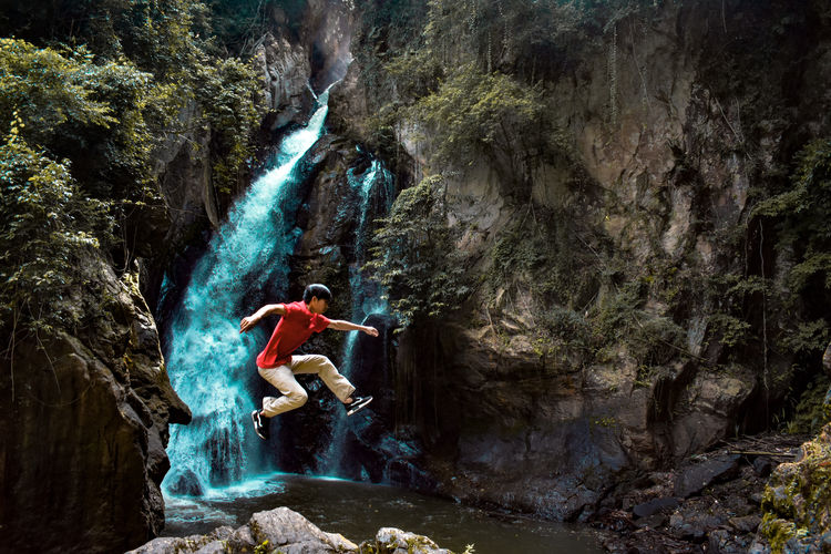 Young man jumping over rock formation against waterfall in forest