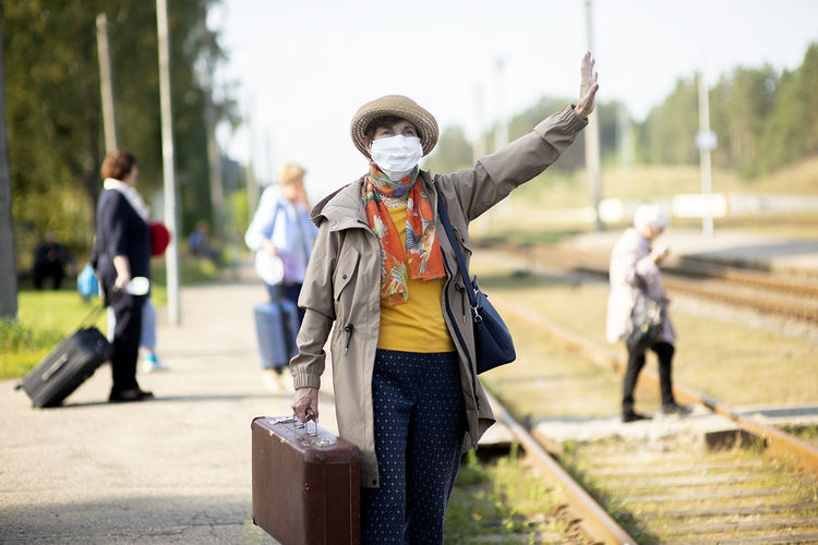 Senior woman wearing flu mask raising hand while standing on railroad station platform