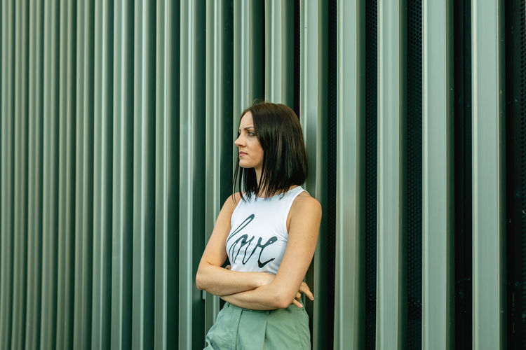 Thoughtful young woman looking away while standing against corrugated iron