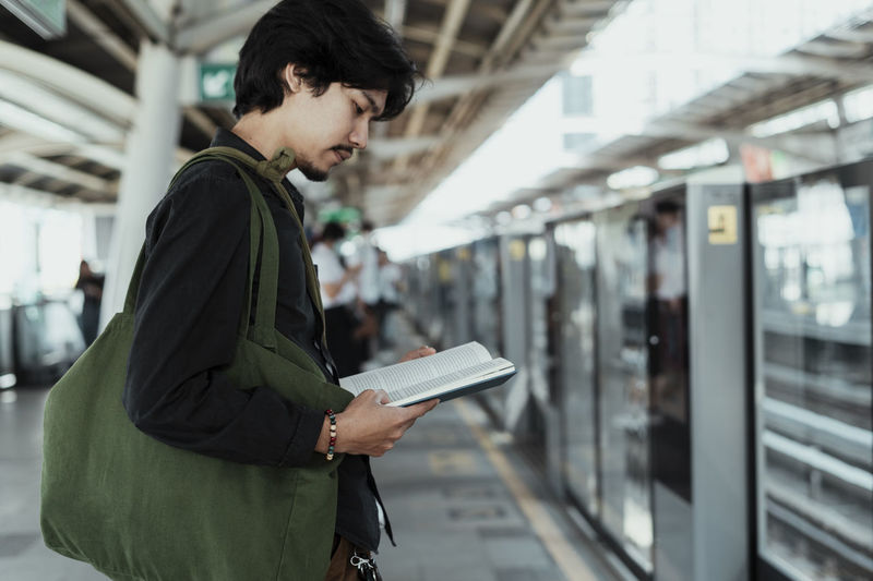 Side view of man reading book while standing at railroad station