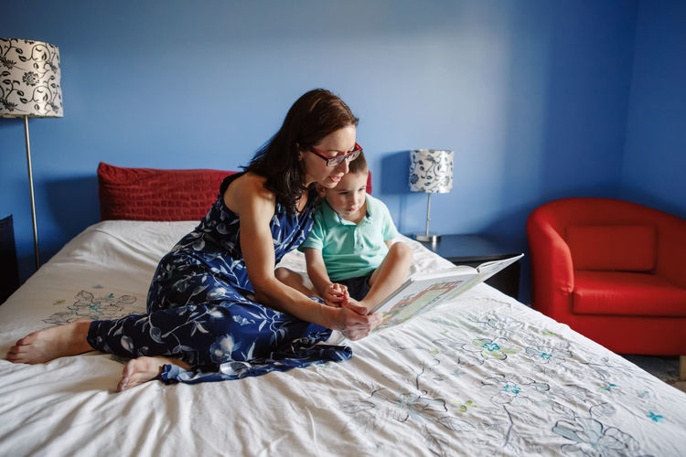 Mother with son boy sitting on bed in bedroom at home and reading book together. child kid early ed