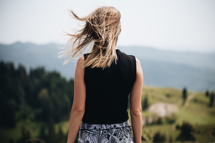 Rear view of woman with tousled blond hair standing against mountains