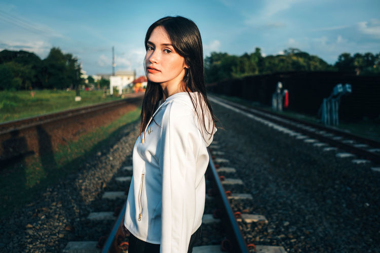 Portrait of young woman standing on railroad track against sky