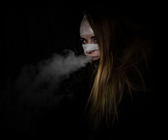Portrait of woman with bandages on face smoking against black background