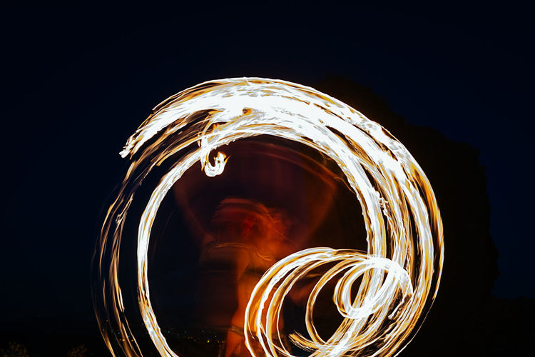Long exposure of unrecognizable skilled fire dancer with fire fan standing on dark street during performance on night time in city