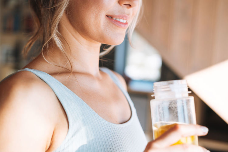 Young fitness blonde woman with bottle of water with lemon doing morning exercises near window 
