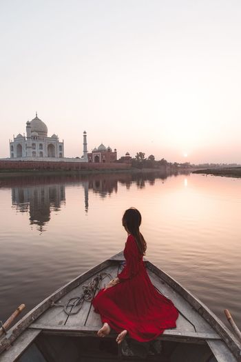Side view of woman wearing red dress sitting on rowboat in lake against sky during sunset
