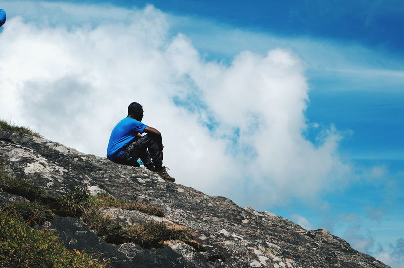 Low angle view of man sitting on cliff against blue sky